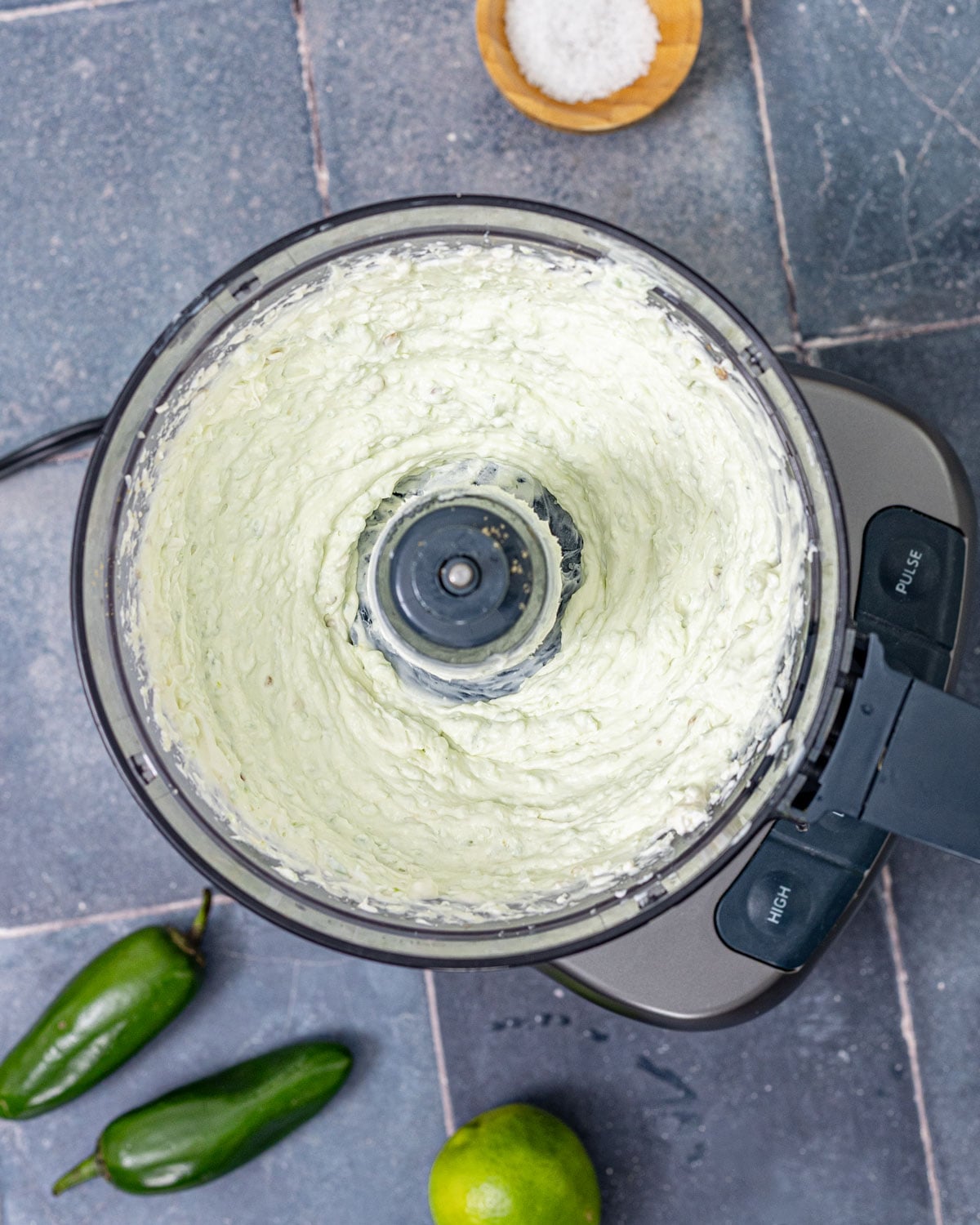 Jalapeño cream cheese blended smooth in food processor with fresh jalapeños and lime in the background
