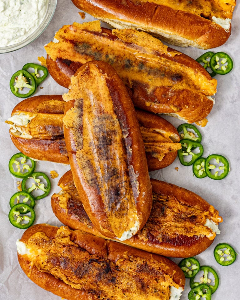 Jalapeño popper hot dogs with seared cheddar crust arranged on parchment paper with sliced jalapeños and a bowl of jalapeño cream cheese