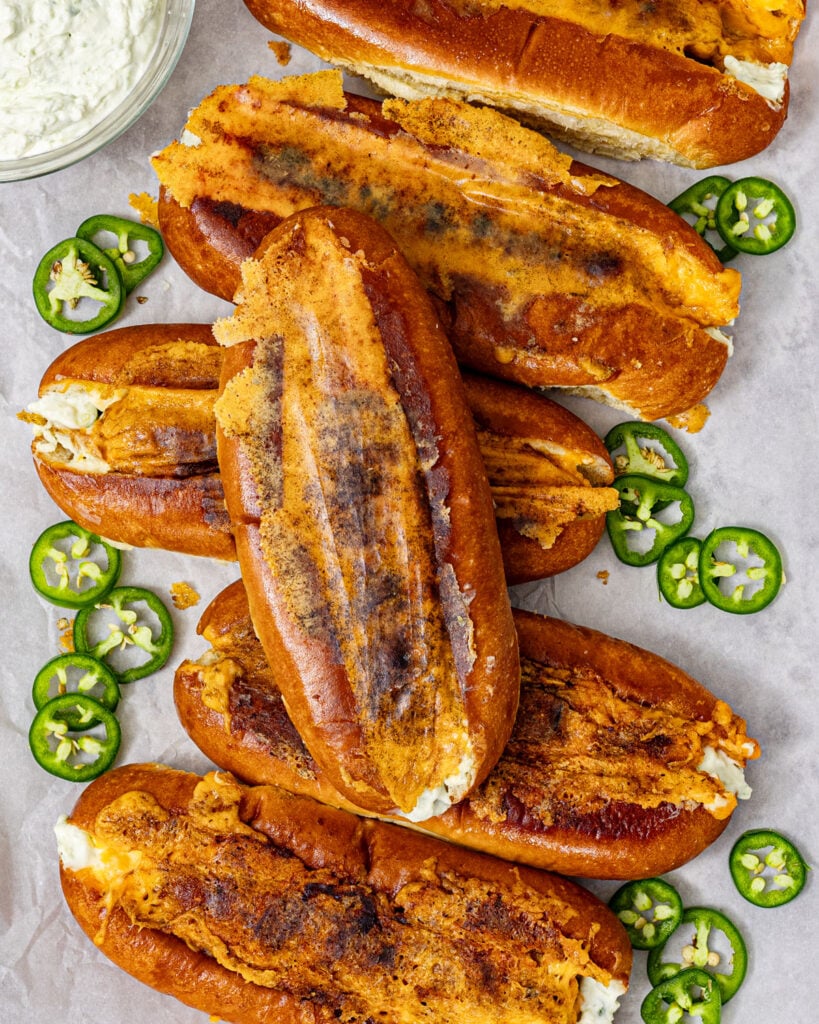 Jalapeño popper hot dogs with seared cheddar crust arranged on parchment paper with sliced jalapeños and a bowl of jalapeño cream cheese