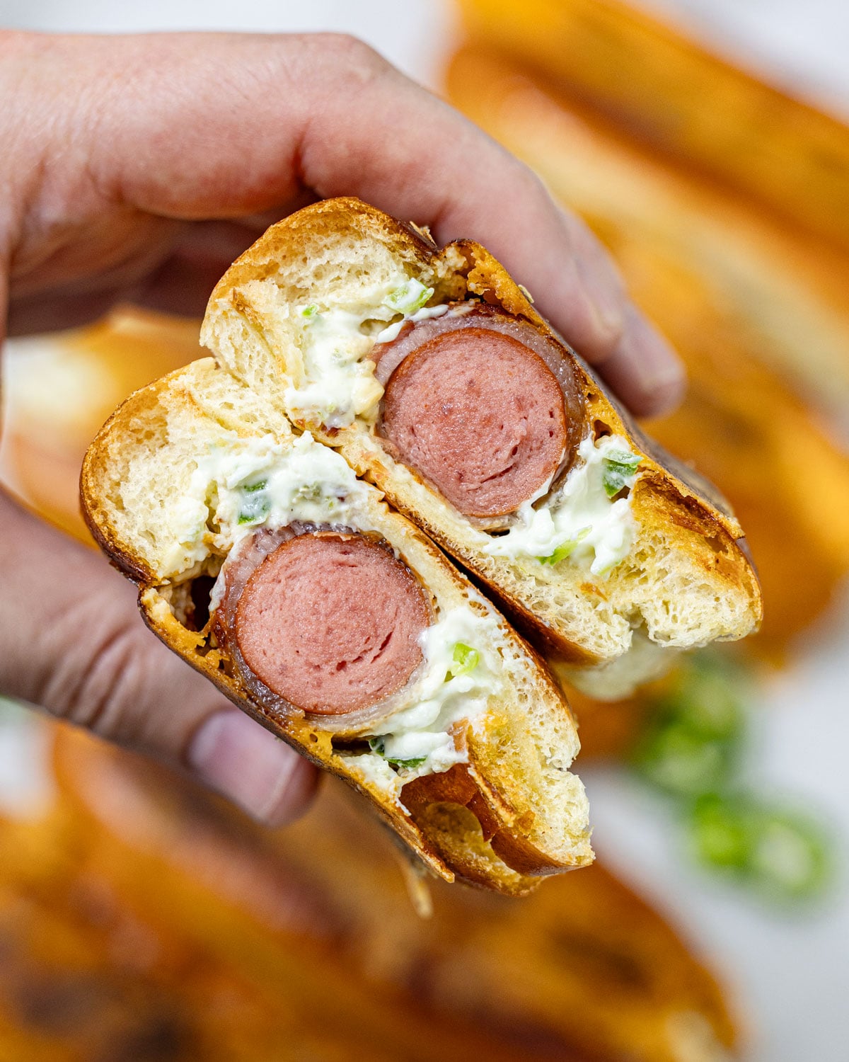 Cross-section of a jalapeño popper hot dog held in two hands showing the bacon-wrapped hot dog surrounded by jalapeño cream cheese