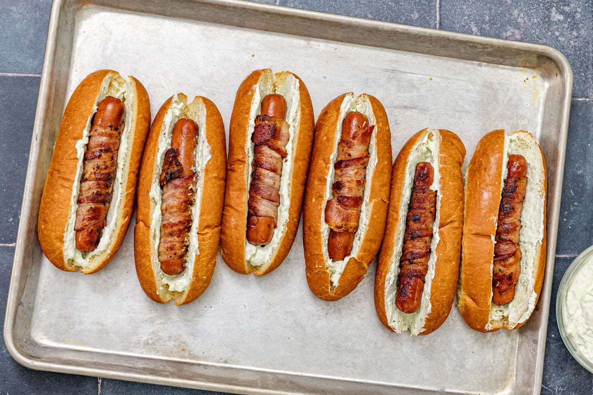 Bacon-wrapped hot dogs loaded with jalapeño cream cheese in brioche buns on a sheet pan, ready for the cheese sear