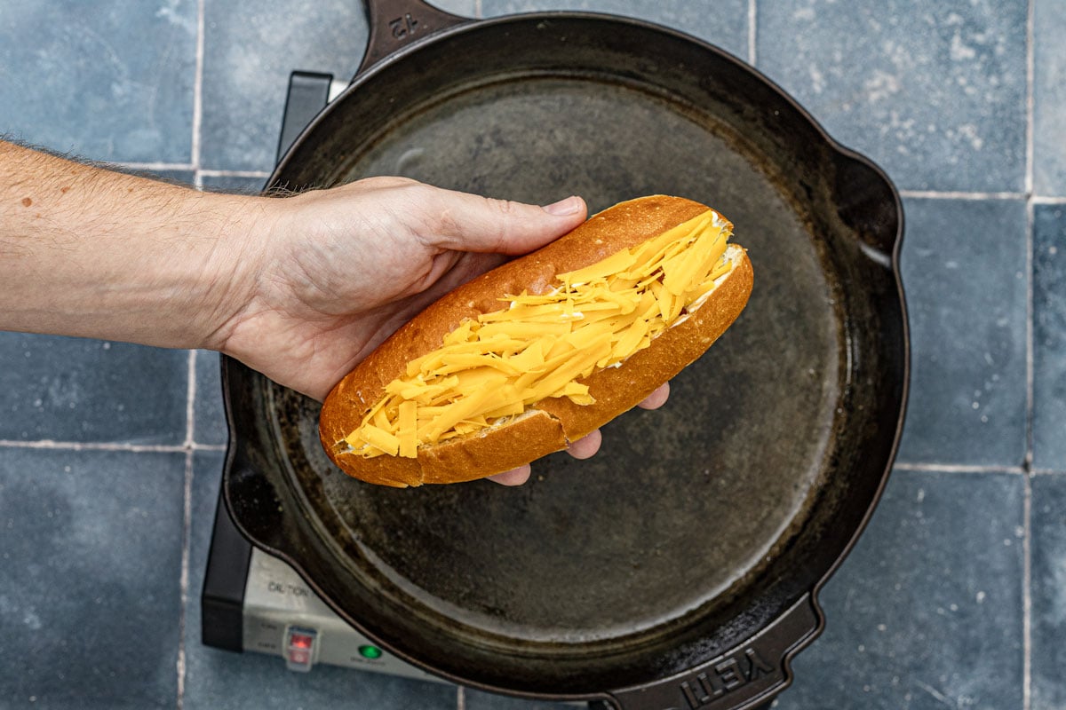 Brioche hot dog bun packed with shredded cheddar held over a cast iron skillet before flipping cheese-side down