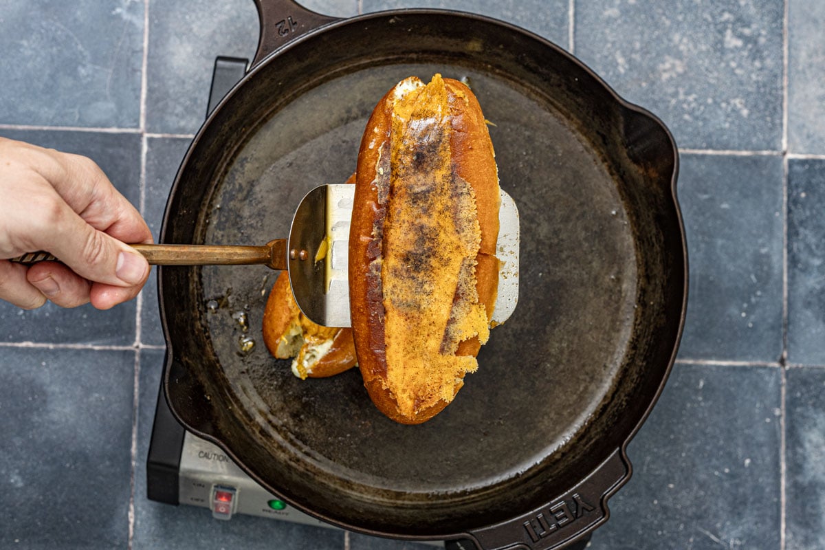 Bench scraper lifting a jalapeño popper hot dog off the cast iron after the cheese crust has seared