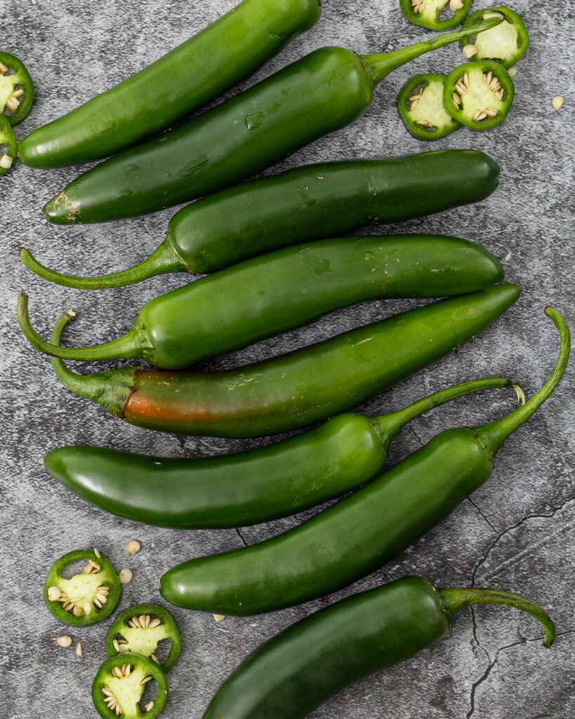 serrano peppers arranged on dark slate with sliced rounds showing seeds, one pepper beginning to ripen