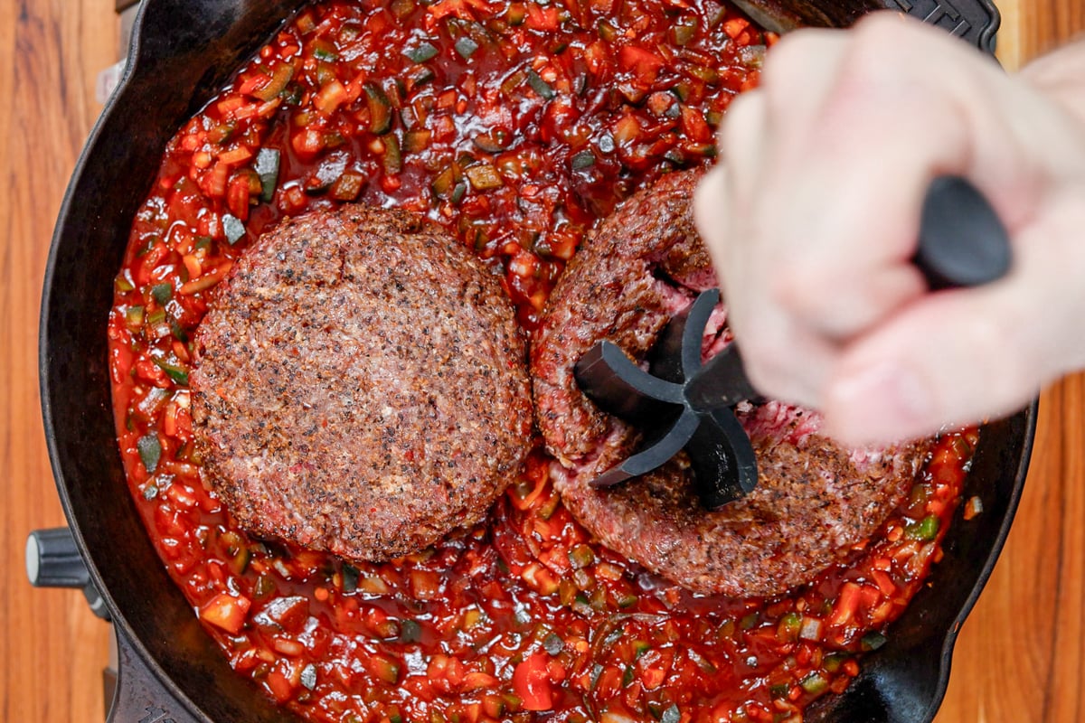 Smoked beef patties being smashed into the simmering sauce with a masher