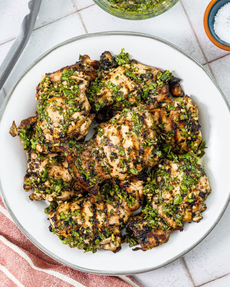 Grilled chicken thighs with char marks topped with fresh chimichurri and a side of sauce