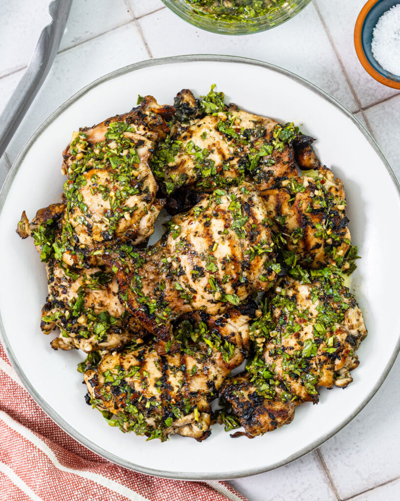 Grilled chicken thighs with char marks topped with fresh chimichurri and a side of sauce