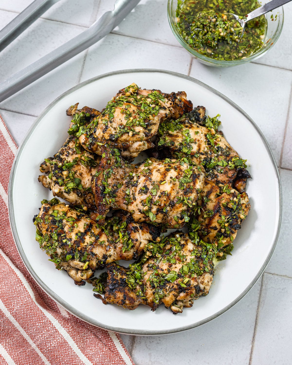 Grilled chimichurri chicken thighs on a white plate topped with fresh chopped chimichurri