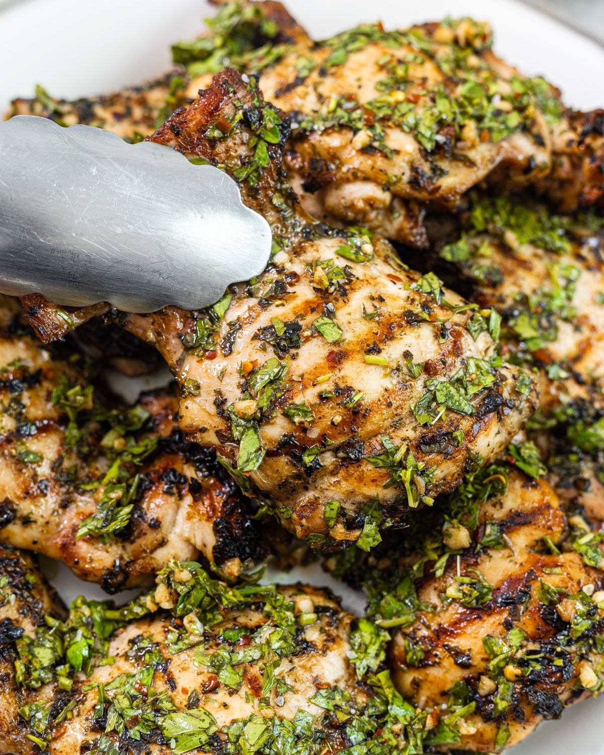 Tongs lifting a charred chimichurri chicken thigh from a stack of grilled thighs