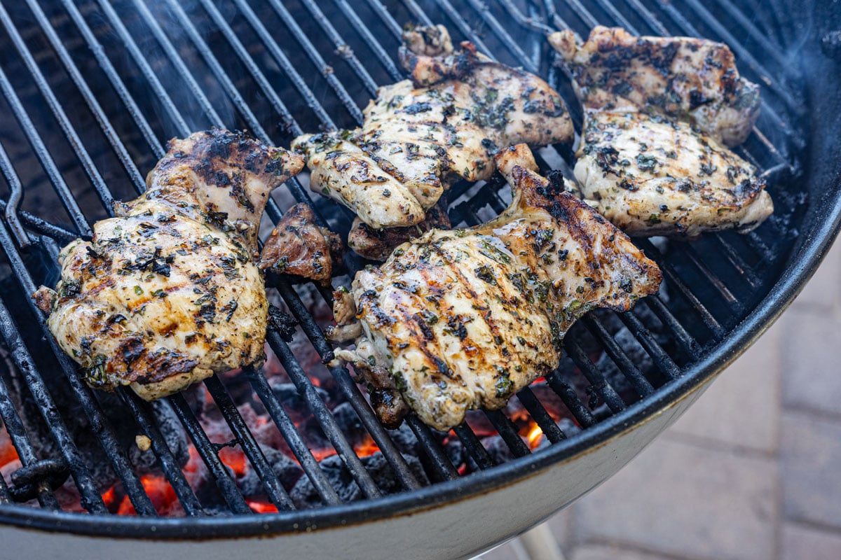 Chimichurri chicken thighs searing over charcoal with grill marks and smoke