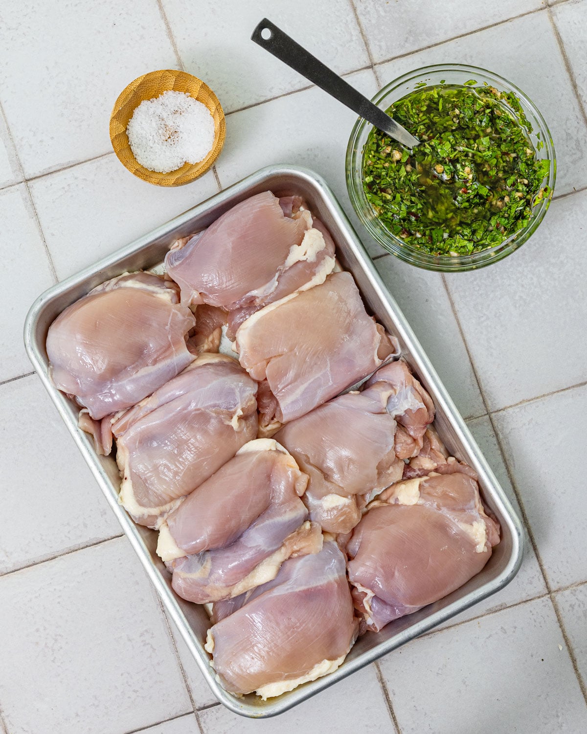 Raw boneless skinless chicken thighs in a metal tray beside small bowls of kosher salt and chimichurri
