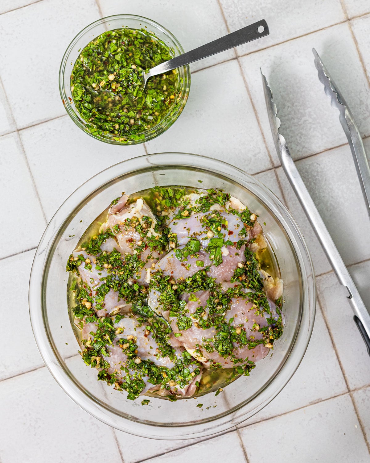 Chicken thighs coated in chimichurri marinade in a glass bowl with a separate reserved bowl of sauce set aside