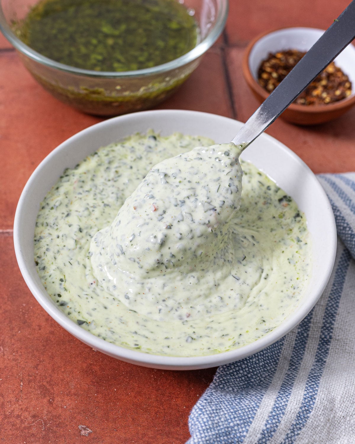 Spoonful of thick chimichurri mayonnaise held above a bowl with red pepper flakes alongside