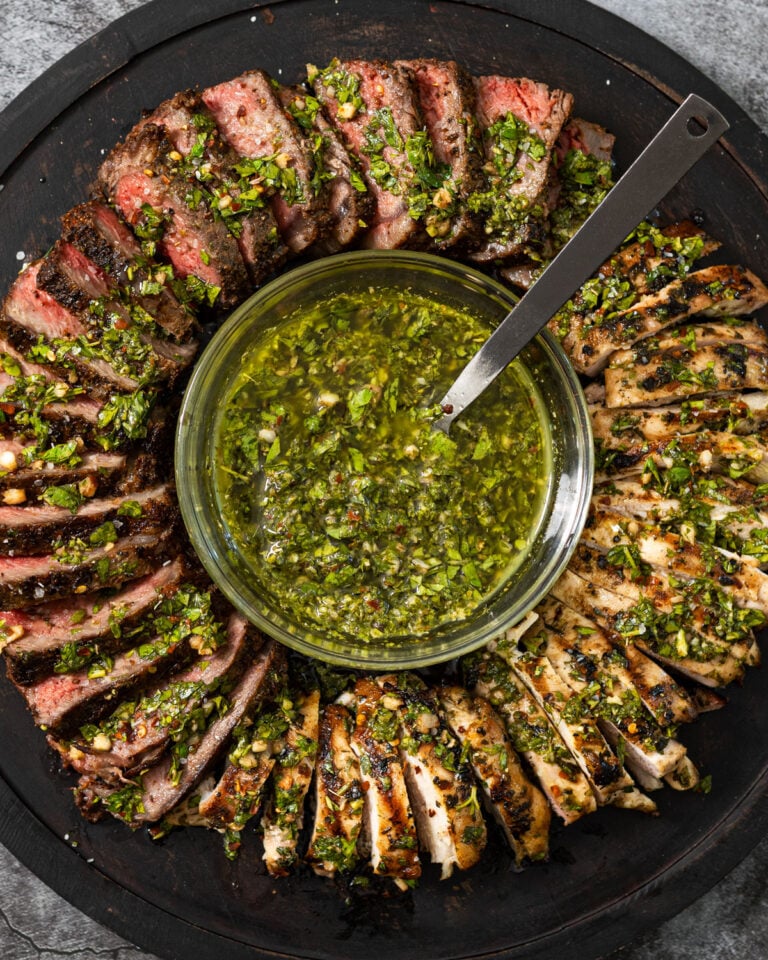 Sliced grilled steak and chicken arranged in a ring around a bowl of chimichurri