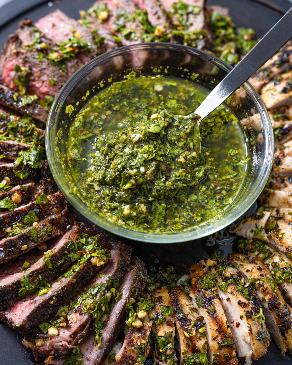 A spoon lifting chunky chimichurri from a bowl set among grilled steak and chicken