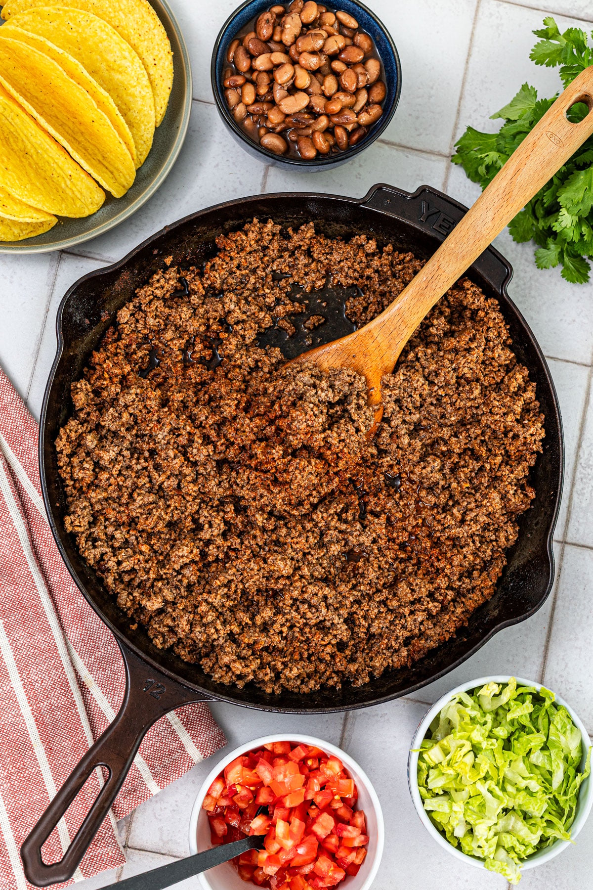 seasoned ground beef taco meat in a cast iron skillet with toppings including beans, shredded lettuce, diced tomatoes, and cilantro