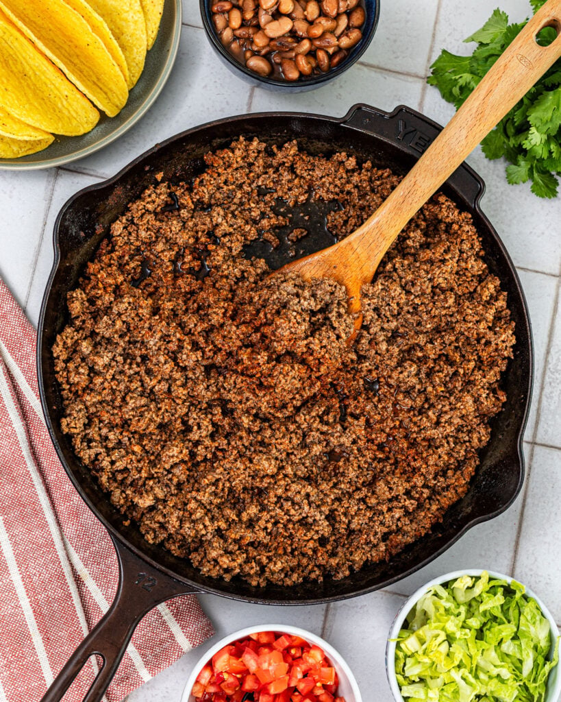seasoned ground beef taco meat in a cast iron skillet with toppings including beans, shredded lettuce, diced tomatoes, and cilantro