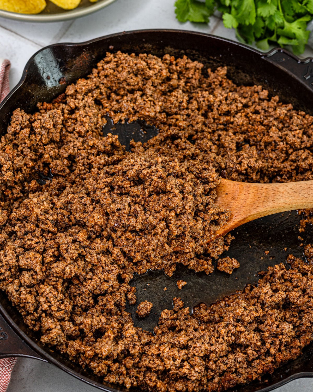 close angle of seasoned ground beef taco meat in a cast iron skillet with wooden spoon scooping, toppings visible in background