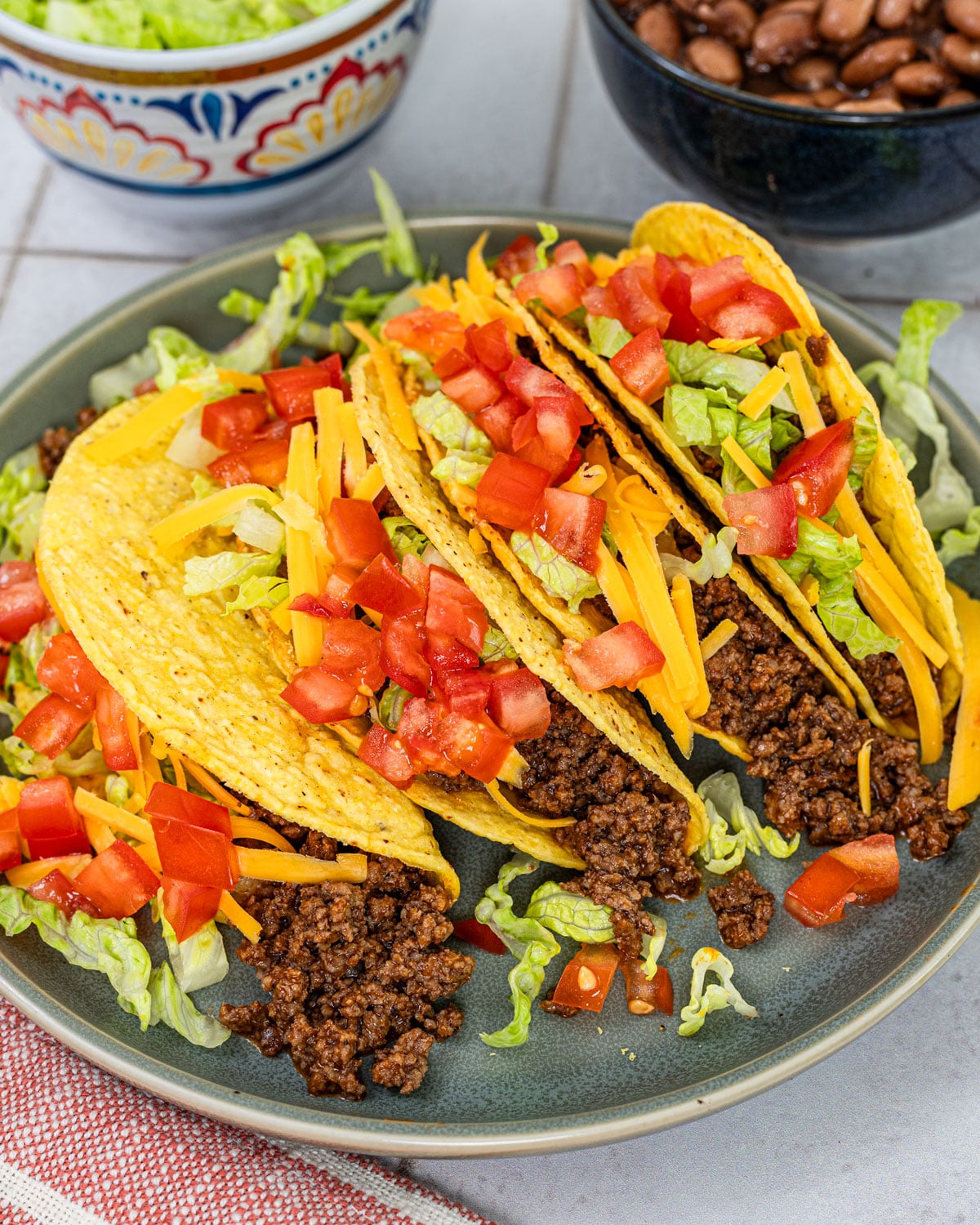 hard shell tacos filled with seasoned ground beef, shredded lettuce, cheddar, and diced tomatoes on a plate with beans and lettuce in background