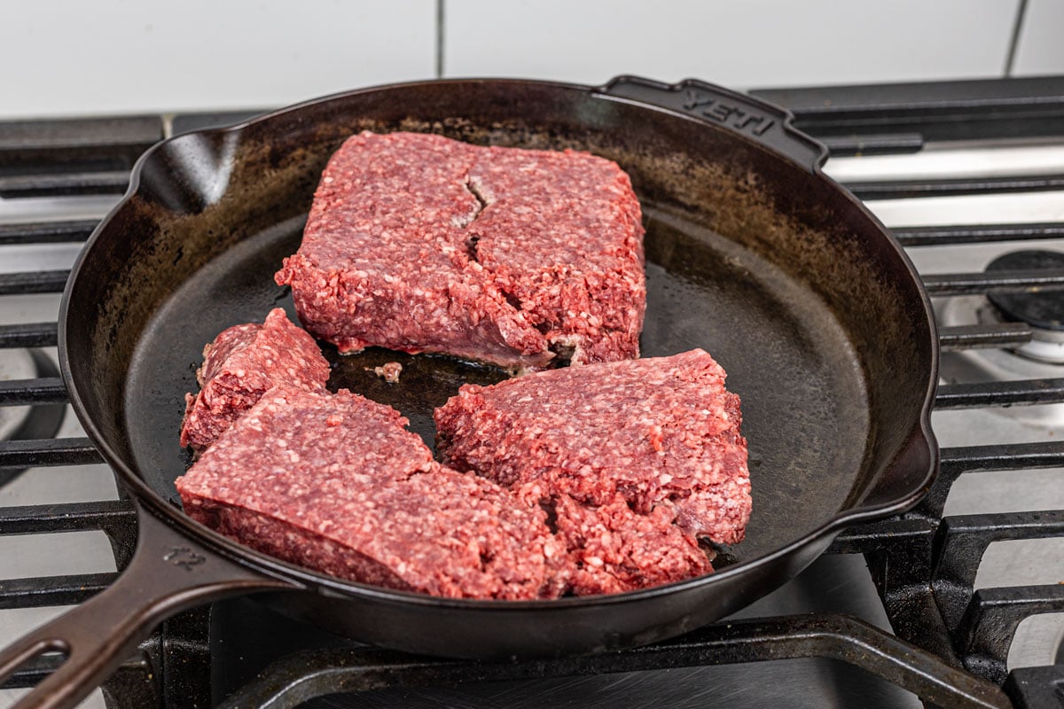raw ground beef pressed flat in a cast iron skillet on a gas stove