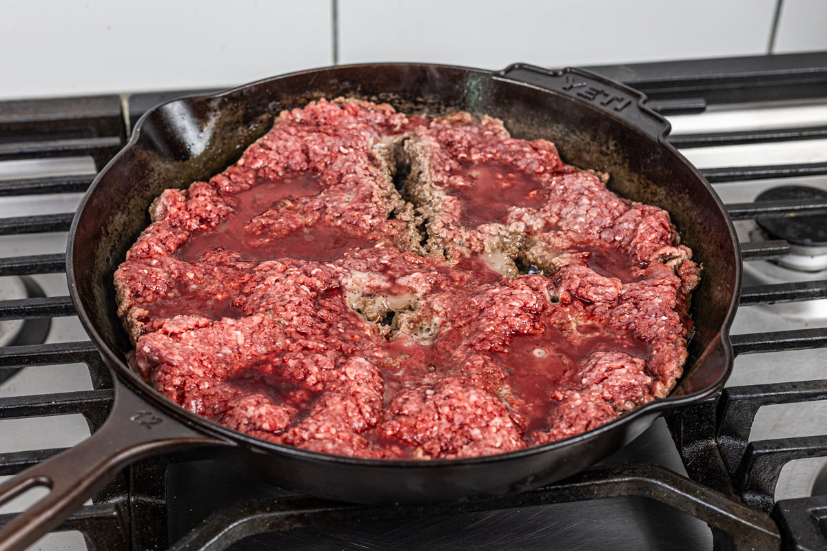 raw ground beef cooking and releasing liquid in a cast iron skillet on a gas stove