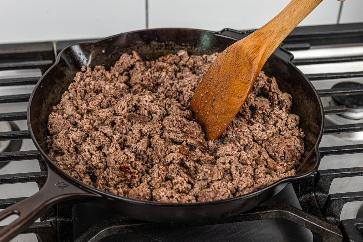 browned ground beef broken into crumbles in a cast iron skillet with wooden spoon