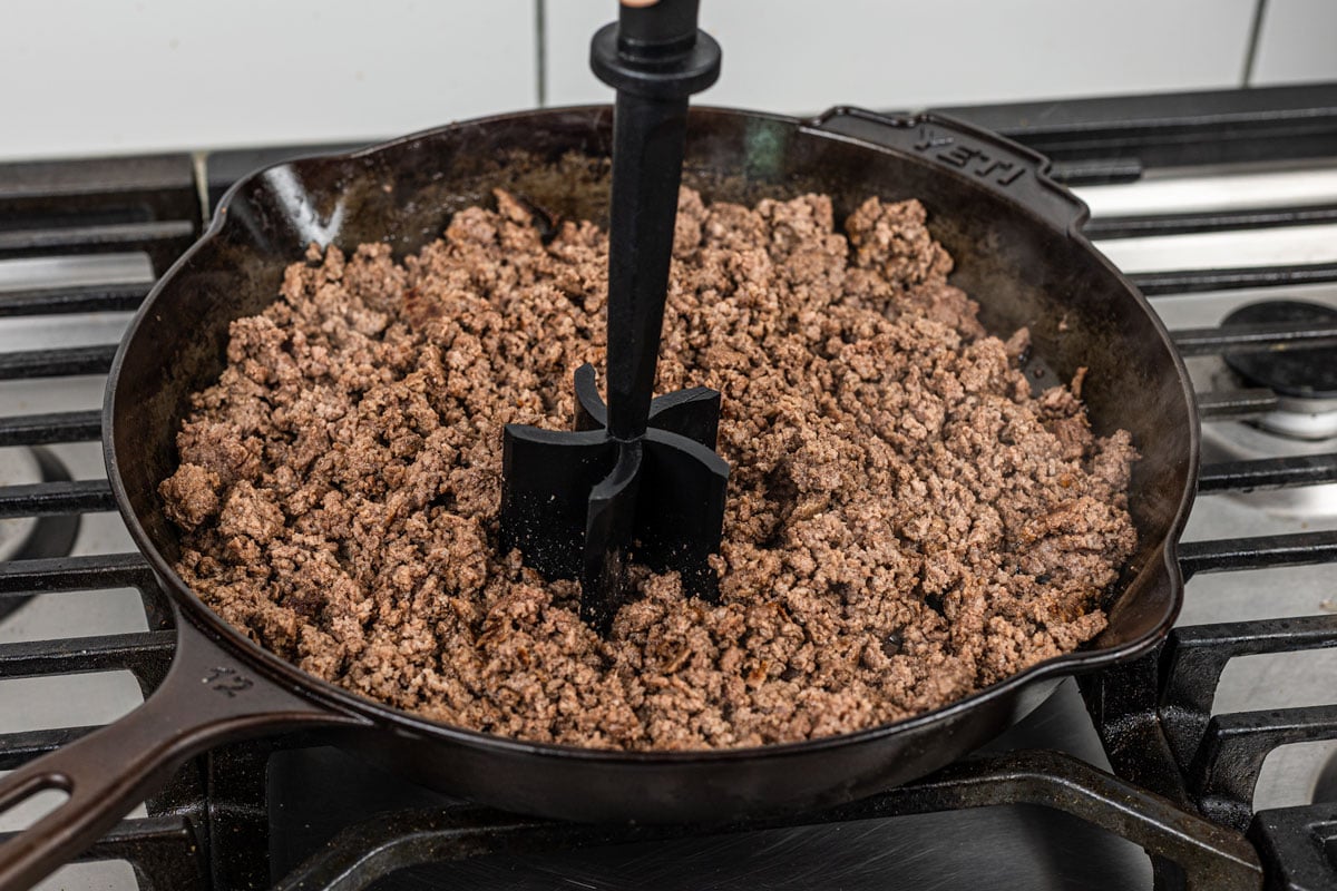 ground beef being broken into fine crumbles with a meat chopper in a cast iron skillet