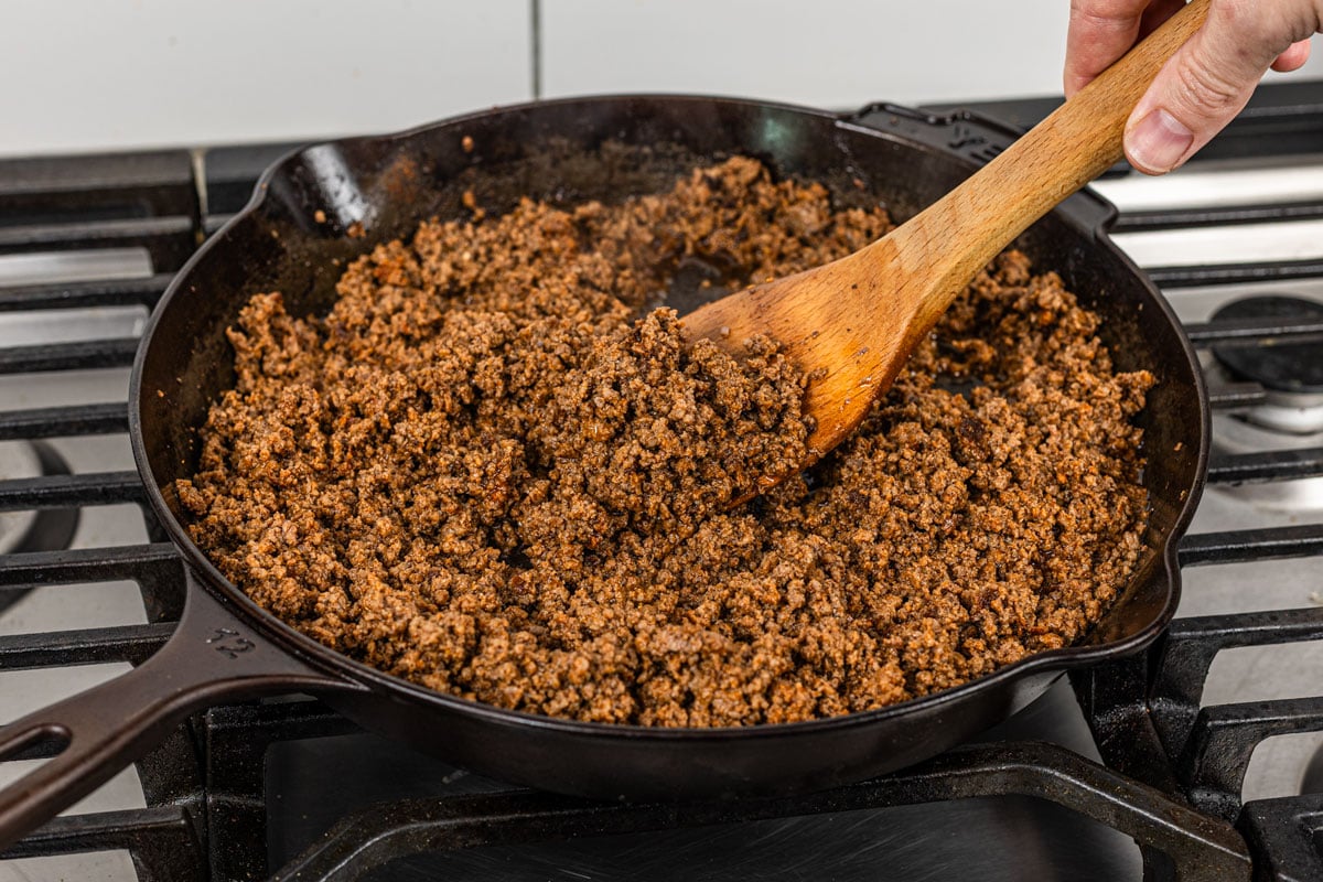 seasoned ground beef being stirred with wooden spoon in cast iron skillet on stove
